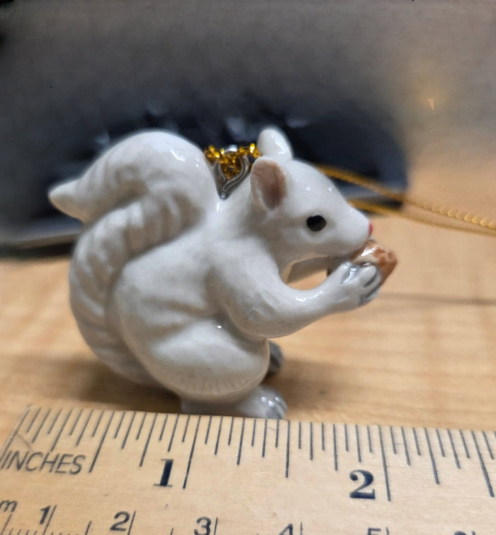 A hand painted porcelain white squirrel ornament with a gold hanging loop, holding an acorn in its mouth close up and size.