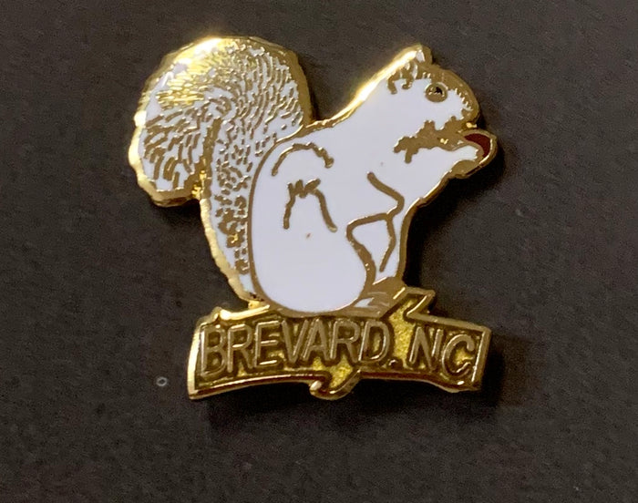 A metal lapel pin in the shape of a white squirrel with gold details, and the text 'BREVARD, NC' on its base.