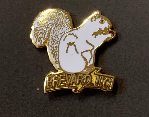 A metal lapel pin in the shape of a white squirrel with gold details, and the text 'BREVARD, NC' on its base.