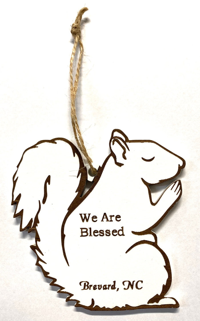 A wooden laser-cut ornament in the shape of a white squirrel praying, with the text 'We Are Blessed, Brevard, NC' engraved on it, hanging by a string.
