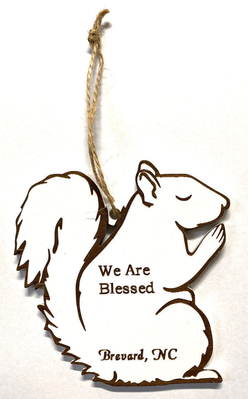 A wooden laser-cut ornament in the shape of a white squirrel praying, with the text 'We Are Blessed, Brevard, NC' engraved on it, hanging by a string.